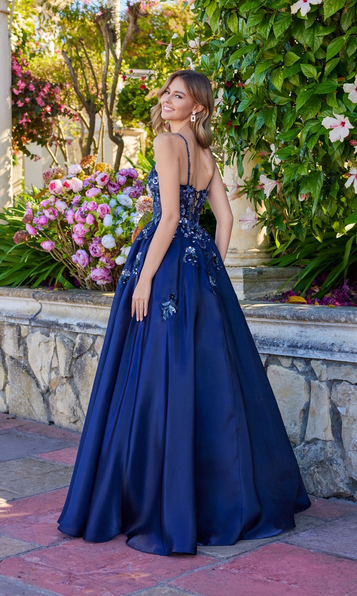 A woman in the Juliet JT2617U long prom dress, featuring navy blue fabric and floral sequin details, stands outdoors by a stone wall and blooming flowers, facing away and smiling over her shoulder.